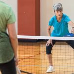 A woman in a teal shirt reaches to return a pickleball shot at the net, with another player facing her in the foreground.
