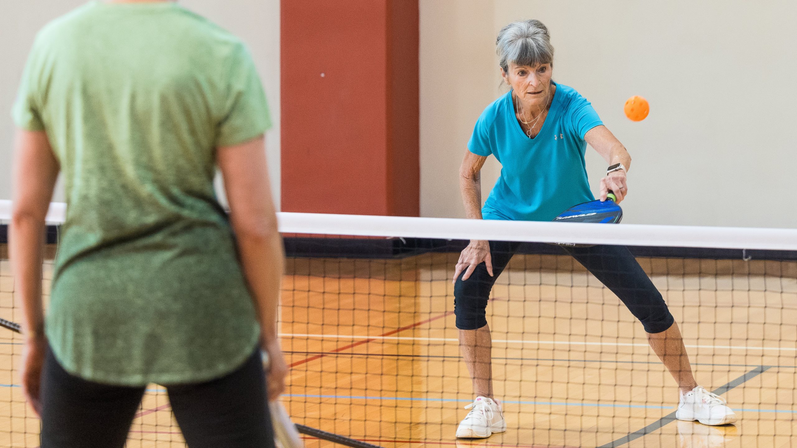 A woman in a teal shirt reaches to return a pickleball shot at the net, with another player facing her in the foreground.