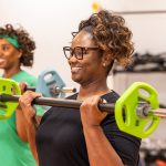 A smiling woman in glasses lifts a barbell loaded with weight plates during a group strength class, with another woman in green lifting behind her.