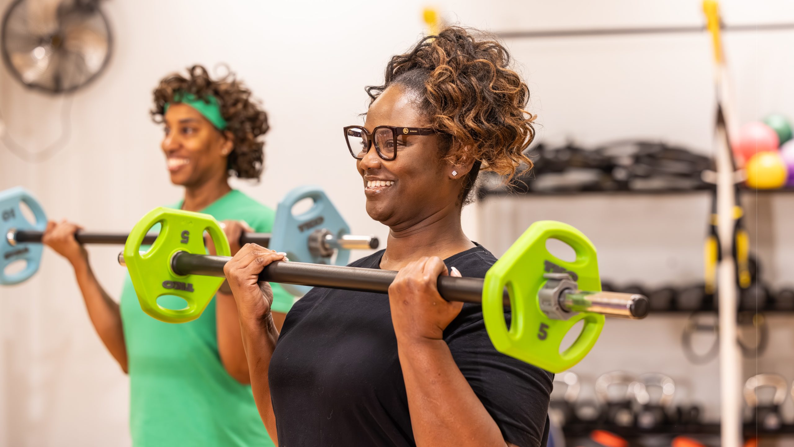 A smiling woman in glasses lifts a barbell loaded with weight plates during a group strength class, with another woman in green lifting behind her.