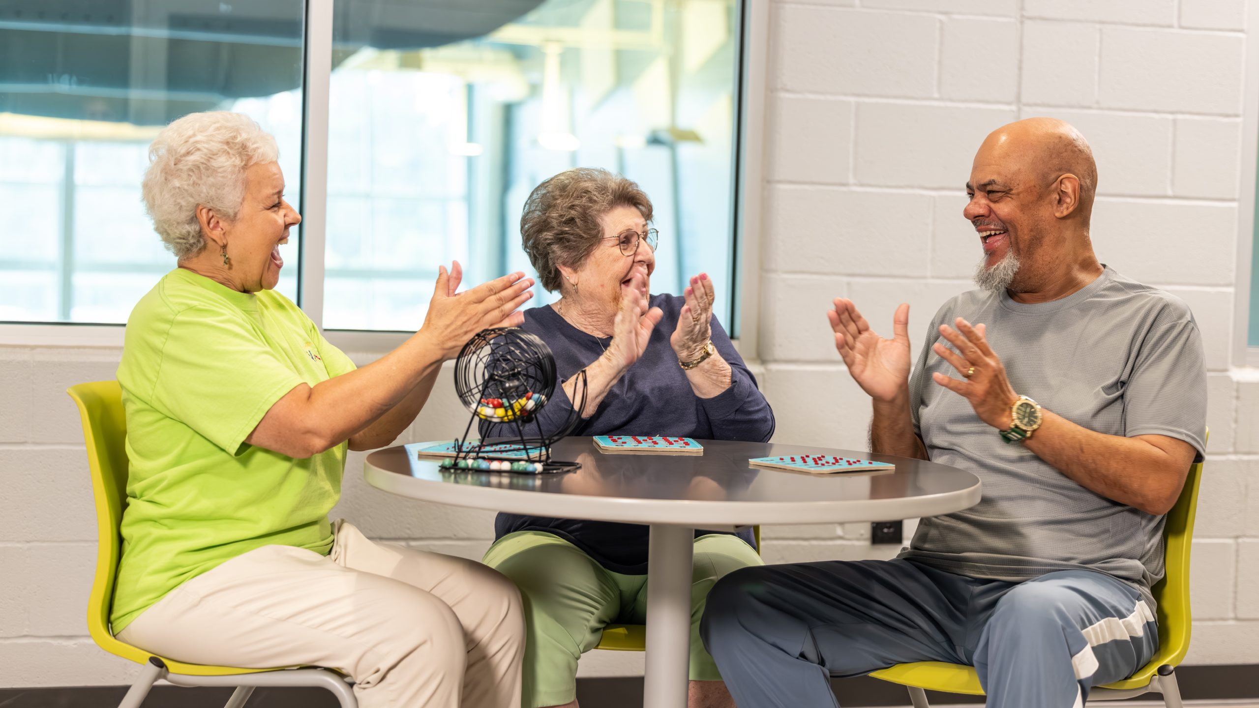 Two women and a man smiling and celebrating together at a round table set with bingo cards and a ball cage during a game.