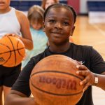 A boy holding a basketball smiles on an indoor court, with two other children holding basketballs behind him.