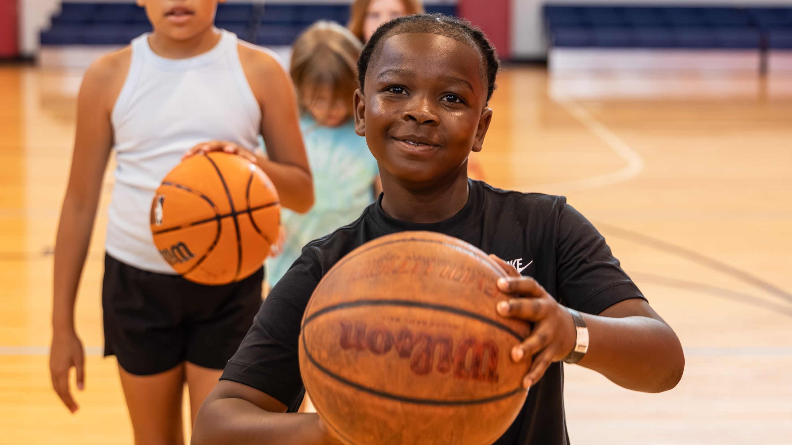A boy holding a basketball smiles on an indoor court, with two other children holding basketballs behind him.