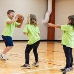Three children play basketball in a gym, with one child holding a basketball while the other two reach toward the ball.