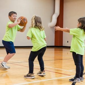 Three children play basketball in a gym, with one child holding a basketball while the other two reach toward the ball.