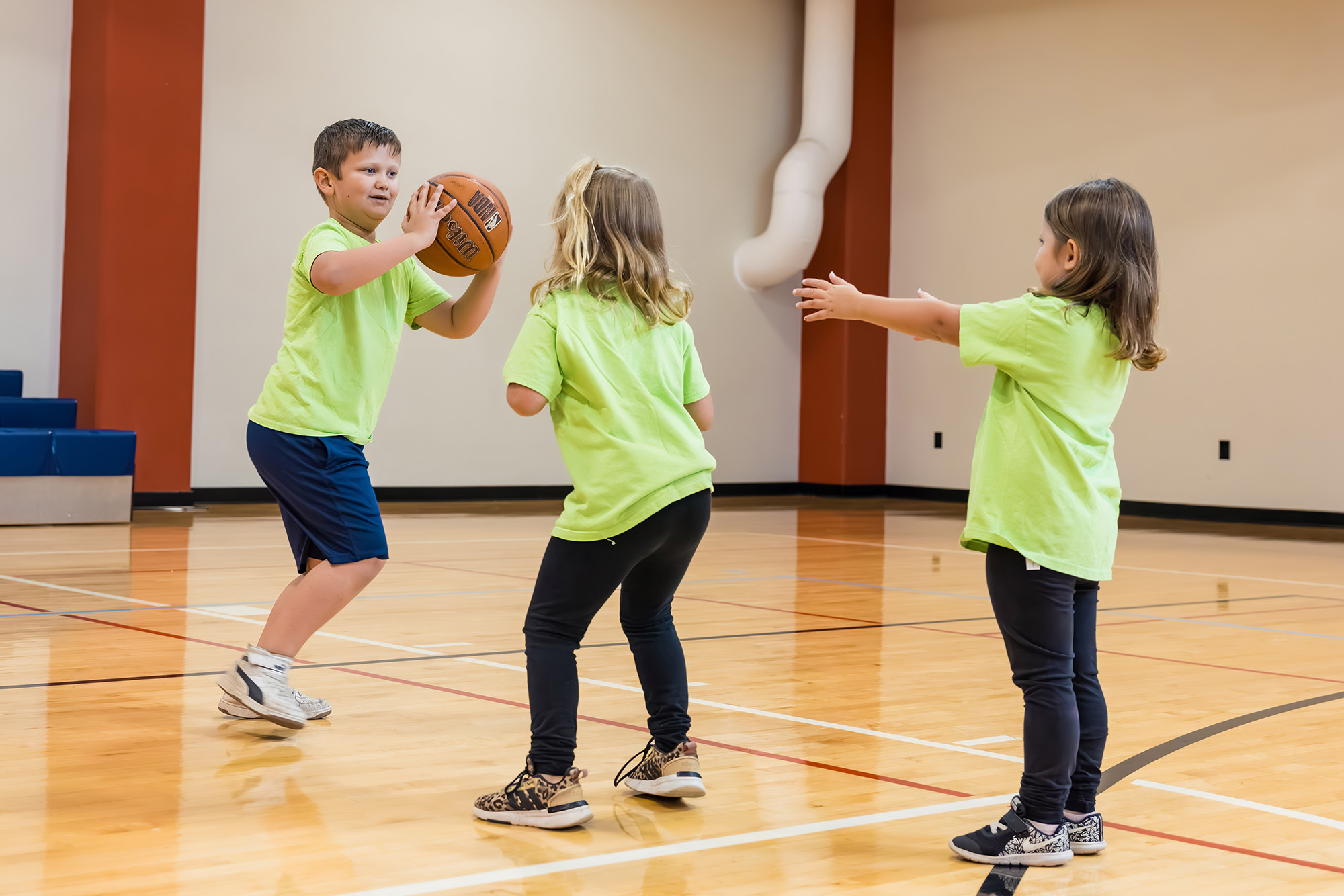 Three children play basketball in a gym, with one child holding a basketball while the other two reach toward the ball.