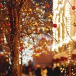 Bare tree branches strung with twinkling string lights and red and gold ornaments, illuminated at night with warmly lit buildings and blurred figures walking in the background.