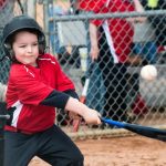 A young child wearing a helmet and red baseball uniform swings a bat at a baseball during a tee-ball game, with spectators and a chain-link fence in the background.