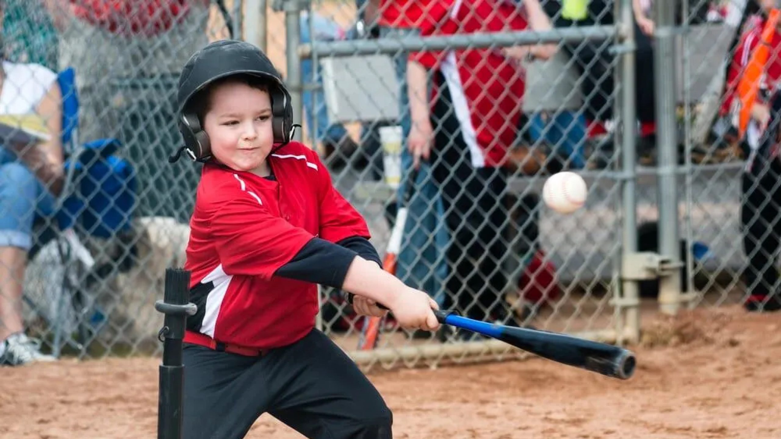 A young child wearing a helmet and red baseball uniform swings a bat at a baseball during a tee-ball game, with spectators and a chain-link fence in the background.