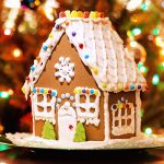 A decorated gingerbread house with white icing snow, colorful candy accents, and gumdrop shingles on the roof, set in front of a blurred, lit Christmas tree.