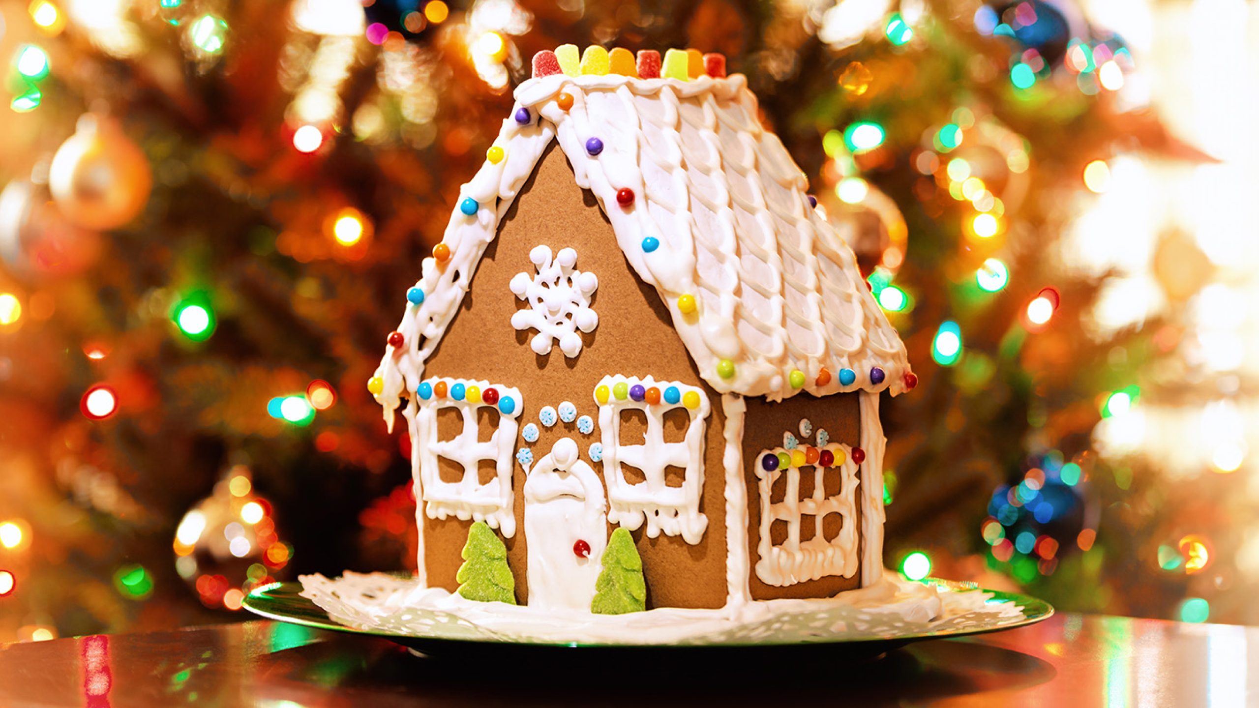 A decorated gingerbread house with white icing snow, colorful candy accents, and gumdrop shingles on the roof, set in front of a blurred, lit Christmas tree.