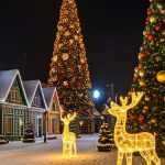 A snowy village street at night lined with festively lit buildings, a tall decorated Christmas tree, illuminated reindeer sculptures, and a smaller ornament-covered tree.