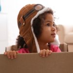 A young girl wearing a brown aviator cap peeks over the edge of a cardboard box, gazing off to the side in a bright, toy-filled room.