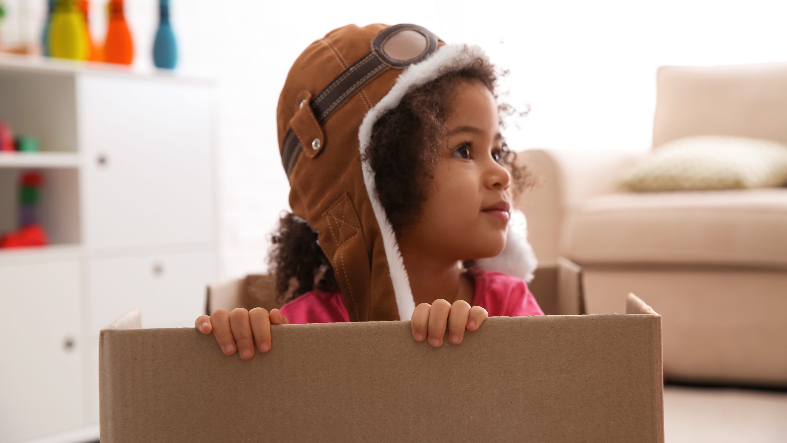 A young girl wearing a brown aviator cap peeks over the edge of a cardboard box, gazing off to the side in a bright, toy-filled room.