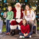 Two boys and a girl smiling alongside Santa Claus, seated in front of decorated Christmas trees and oversized glittering present decorations.