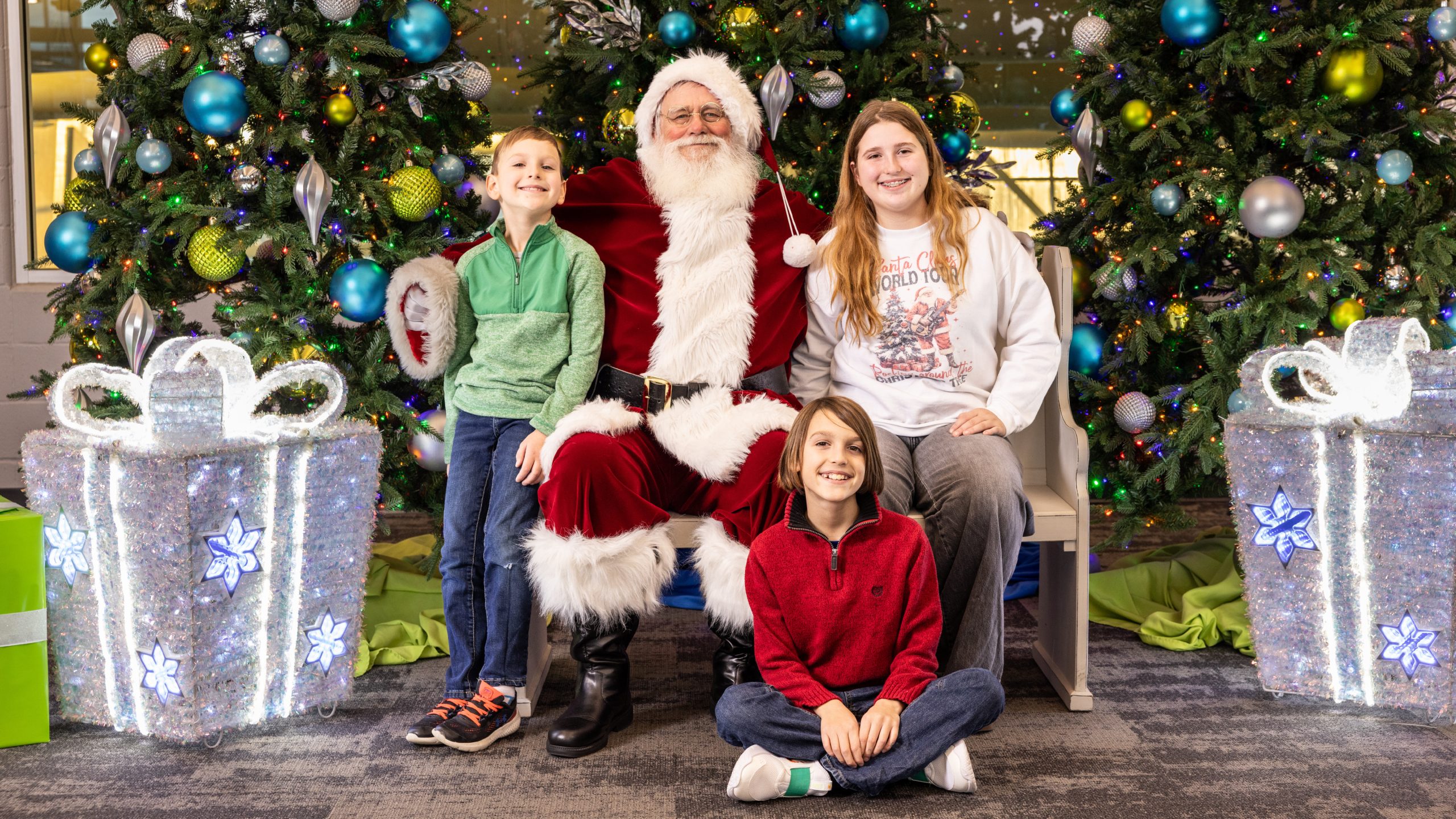 Two boys and a girl smiling alongside Santa Claus, seated in front of decorated Christmas trees and oversized glittering present decorations.