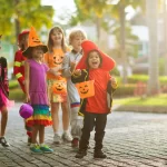 Child in Halloween costume. Mixed race kids and parents trick or treat on street. Little boy and girl with pumpkin lantern and candy bucket. Baby in witch hat. Autumn holiday fun.