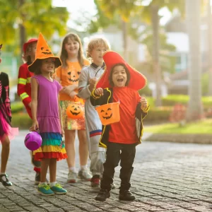 Child in Halloween costume. Mixed race kids and parents trick or treat on street. Little boy and girl with pumpkin lantern and candy bucket. Baby in witch hat. Autumn holiday fun.