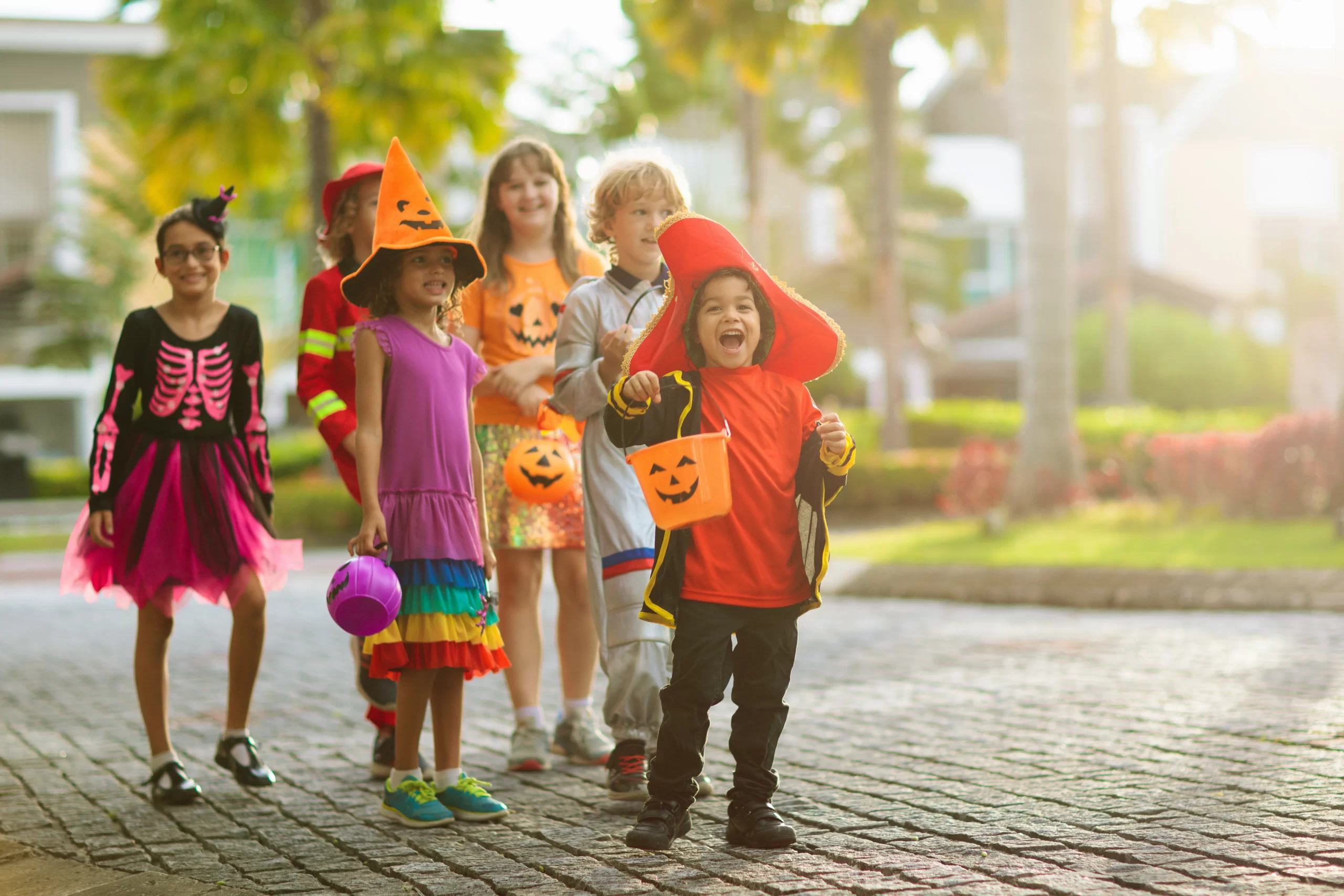 Child in Halloween costume. Mixed race kids and parents trick or treat on street. Little boy and girl with pumpkin lantern and candy bucket. Baby in witch hat. Autumn holiday fun.